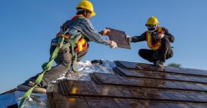 Two roofers in yellow helmets and safety vests installing shingles on a roof.